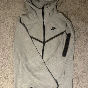 Grå NikeTech Hoodie (Xs) - Säljer en grå hoodiejacka från Nike med svarta detaljer och dragkedja framtill. Jackan har en ficka med dragkedja på ärmen och en sportig design med huva. Perfekt för dig som gillar stilren och bekväm streetwear.