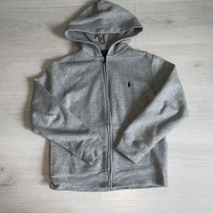 Grå hoodie från Polo Ralph Lauren - Säljer en klassisk grå hoodie från Polo Ralph Lauren med dragkedja och huva. Tröjan har broderad logga på bröstet och är tillverkad i mjuk bomull. Perfekt för en avslappnad stil.