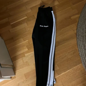 Svarta träningsbyxor från Palm Angels med vita ränder - Svarta träningsbyxor från Palm Angels med två vita ränder längs sidorna och logga på låret. Byxorna har resår i midjan och är raka i modellen, perfekta för en sportig och trendig look.
