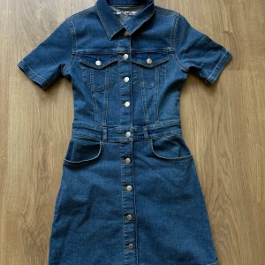 Jeansklänning med kort ärm och knappar - Snygg jeansklänning i blå denim med korta ärmar, krage och knäppning framtill. Klänningen har fickor både fram och bak samt markerad midja för en klassisk look. Perfekt för dig som gillar denim och vill ha en cool vardagsstil.