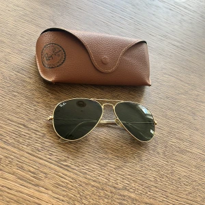 Ray-Ban Aviator solglasögon med guldiga bågar - Klassiska Ray-Ban Aviator solglasögon med tunna guldiga metallbågar och mörkgröna glas. Kommer med original brunt fodral i läderlook. Perfekt accessoar för soliga dagar.