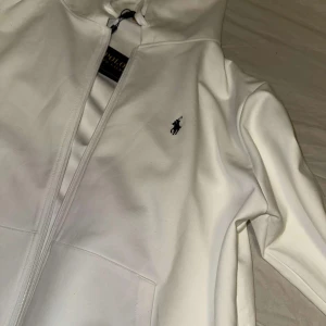 Vit hoodie från Ralph Lauren - Säljer en stilren vit hoodie från Ralph Lauren med dragkedja och det klassiska logotypbroderiet på bröstet. Perfekt för en avslappnad och snygg look. Hör av dig vid intresse! Storlek M-L