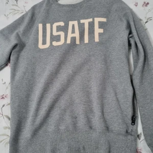 Nike USATF - Grå sweatshirt från Nike med USATF-tryck framtill.. Mjuk insida och ribbade muddar. Perfekt för en sportig look.                                  knappt använd.