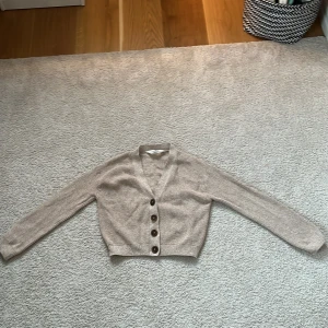 Beige stickad kofta med knappar - Säljer en beige stickad kofta med v-ringning och stora bruna knappar framtill. Koftan är croppad och har långa ärmar. Perfekt för lager-på-lager och enkel att matcha med olika outfits.