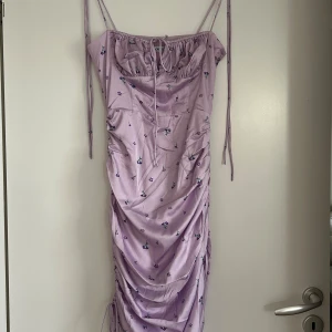 Ljuslila blommig klänning från Adoore - Supersöt ljuslila klänning från Adoore med små blommor, smala axelband och rynkade detaljer längs sidorna. Klänningen har knytning framtill och en figurnära passform som framhäver siluetten. Perfekt för sommarens alla tillfällen! Aldrig använd