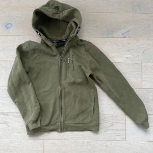 Olivgrön hoodie från Sail Racing - Säljer en olivgrön hoodie från Sail Racing med dragkedja framtill och på bröstfickan. Jackan har huva med justerbar snörning och mjukt fleecefoder på insidan. Broderad logga på bröstet och ribbade muddar vid ärmslut och nederkant.