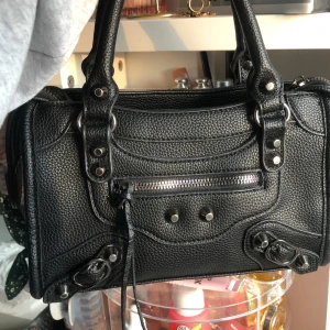 Svart handväska i skinn med detaljer - Säljer en svart handväska i skinn med silverfärgade metalldetaljer och dragkedja framtill. Väskan har två rejäla handtag och en strukturerad yta. Perfekt för dig som vill ha en stilren och cool accessoar till din outfit.