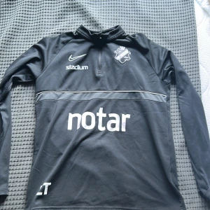 Svart långärmad träningströja från Nike AIK - Svart långärmad träningströja från Nike med AIK-logga på bröstet och flera sponsorer tryckta fram och bak. Tröjan har halv dragkedja framtill och är tillverkad i Dri-FIT-material som andas. Perfekt för fotbollsträning.