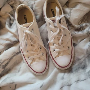 Vita Converse All Star sneakers - Klassiska vita Converse All Star sneakers med lågt skaft och snörning. Skorna har rund tå, platt sula och den ikoniska röda randen längs sulan. Perfekta för en avslappnad och tidlös stil.