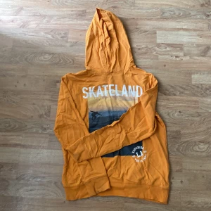 Orange hoodie med SKATELAND-tryck - Säljer en orange hoodie med trycket 'SKATELAND' och en bild på en skateboardåkare på ryggen. Tröjan har huva och långa ärmar, samt en vit detalj med text på nedre delen av ryggen. Perfekt för dig som gillar streetwear.