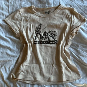 Beige t-shirt med Oregon-tryck från Brandy Melville - Säljer en beige t-shirt från John Galt med ett svart tryck av en man och en björn samt texten 'OREGON' på framsidan. T-shirten har rund halsringning och är tillverkad i 100% bomull. Perfekt för en avslappnad stil.