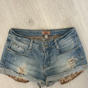 Ljusblå low waist jeansshorts med slitningar från Bershka - Säljer ett par ljusblå jeansshorts från Bershka med slitna detaljer och fransar nertill. Shortsen har klassisk femficksdesign och är dekorerade med glittriga paljetter på insidan av bensluten.