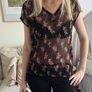 Svart transparent blus med blommigt mönster - Säljer en svart transparent blus med blommigt mönster i beige och rosa toner. Blusen har korta ärmar och en lös passform.