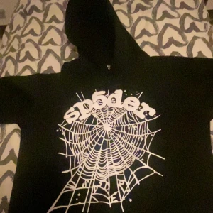 Svart hoodie med spindelnätsmotiv från SP5DER - Säljer en svart hoodie från SP5DER med stort vitt spindelnätsmönster och texten 'sp5der' på framsidan. Tröjan har huva och långa ärmar, perfekt för dig som gillar streetwear och unika tryck.