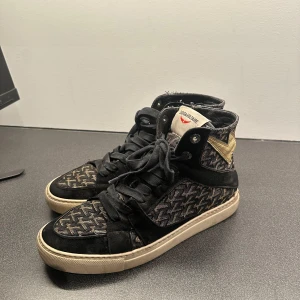 Svarta sneakers från Zadig & Voltaire med guld-detalj - Snygga svarta high-top sneakers från Zadig & Voltaire med mönstrad textil, svarta mockadetaljer och en guldig blixt på sidan. Skorna har snörning och vit platt sula. Perfekta för dig som vill sticka ut med en cool streetstil. 