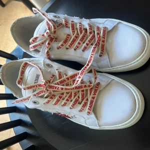 Vita sneakers från Zadig & Voltaire - Säljer ett par vita sneakers från Zadig & Voltaire med röda detaljer och text på skosnörena. Skorna har rund tå, platt sula och är tillverkade i canvas med gummisula. Perfekta för en avslappnad och trendig stil.