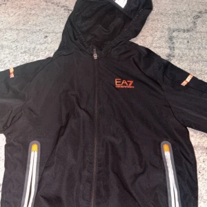 Svart vindjacka från Emporio Armani EA7 - Säljer en svart vindjacka från Emporio Armani EA7 med huva och dragkedja framtill. Jackan har orange EA7-logga på bröstet och detaljer vid fickorna. Reflekterande detaljer och sportig look.