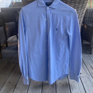 Blå skjorta från Polo Ralph Lauren - Säljer en klassisk ljusblå skjorta från Polo Ralph Lauren i modellen Knit Oxford. Skjortan har lång ärm, knappar framtill och den ikoniska broderade loggan på bröstet. Perfekt till sommaren. Nästan helt oanvänd så ser helt ny ut, pris kan diskuteras. 