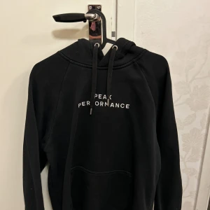 Peak Performance hoodie - Svart hoodie från Peak Performance med vit text på bröstet. Tröjan har huva med dragsko och en stor magficka framtill. Perfekt för en avslappnad stil i storlek M.