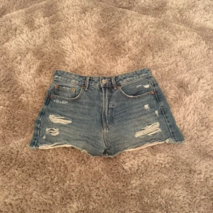 Blå jeansshorts från Stradivarius - Säljer ett par blå jeansshorts från Stradivarius i storlek 38 (M/W28). De har slitna detaljer och fransig kant nertill. Klassisk modell med knapp och dragkedja framtill.