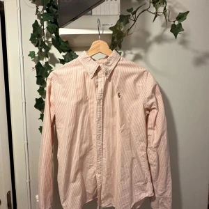 Laxrosa och vit randig skjorta från Ralph Lauren - Säljer en klassisk laxrosa och vit randig skjorta från Ralph Lauren i slim fit-modell. Skjortan har knappar framtill, krage och den ikoniska broderade loggan på bröstet. Perfekt för en stilren look. Sitter som en S 