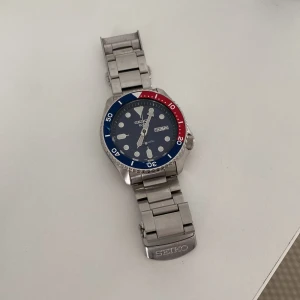 Seiko 5 Sports automatisk herrklocka - Klassisk Seiko 5 Sports automatisk klocka med blå urtavla, röd och blå vridring samt robust länkarmband i silverfärgat stål. Urtavlan har tydliga index och dag/datum-visning. Perfekt för dig som gillar sportig stil och mekaniska urverk. Skriv om du har frågor, priset är diskuterbart och kan går ner i pris vid snabb affär