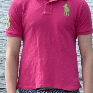 Rosa pikétröja från Polo Ralph Lauren - Säljer en klassisk rosa pikétröja från Polo Ralph Lauren med grönt broderat logotyp på bröstet och siffra på ärmen. Tröjan har korta ärmar, krage och knappar framtill. Tillverkad i 100% bomull.