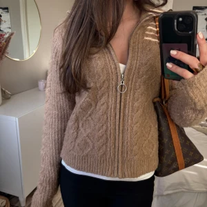 Beige stickad kofta med dragkedja - Säljer en beige stickad kofta med dragkedja och diskret mönster. Koftan har lång ärm och ribbade muddar. Perfekt för lager-på-lager och passar till det mesta.