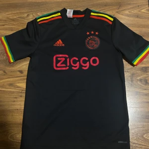 Ajax svart bortatröja 2021/22 från Adidas - Säljer en svart Ajax bortatröja från Adidas med röd Ziggo-logga och klubbmärke. Tröjan har korta ärmar med röd, gul och grön detalj samt Adidas-ränder på axlarna. Materialet är lätt och andas, perfekt för fotboll eller streetwear. Tröjan har ett märke på magen efter att en tråd dragits ur. Man kan se märket om man kollar noga på första bilden! 