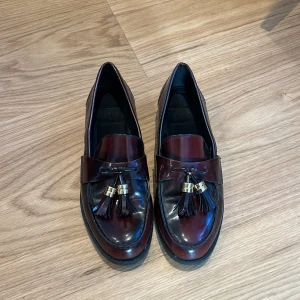 Vinröda loafers med tofsar och gulddetaljer - Säljer ett par  vinröda loafers i fejkskinn med dekorativa tofsar och guldfärgade detaljer framtill. Skorna har rund tå och platt sula. Använda max 3 gånger.