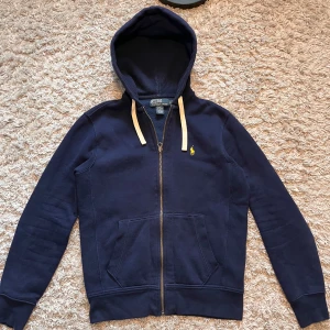 Marinblå hoodie från Polo Ralph Lauren - Säljer en marinblå hoodie från Polo Ralph Lauren med dragkedja och huva. Tröjan har gul broderad logga på bröstet och vita snören vid huvan. Tillverkad i bomull och polyester, perfekt för en avslappnad stil.