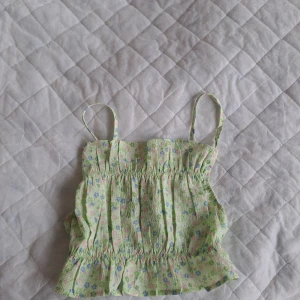 Grön blommig linnetopp med volang - Söt ljusgrön linnetopp med små blommor i rosa, blått och vitt. Toppen har smala axelband, smockad överdel och volang nertill. Perfekt för varma dagar och somriga outfits.