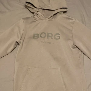 Björn Borg hoodie - Säljer en beige/grå hoodie från Björn Borg med trycket 'BORG SINCE 1956' på bröstet. Tröjan har huva och en magficka framtill. Perfekt för en avslappnad stil och enkel att matcha med det mesta. Tröjan är lite större i storlek så den passar er som har xs/s och är bara använd fåtal gånger. Pris kan diskuteras. Bara höra av er vid minsta fundering😊