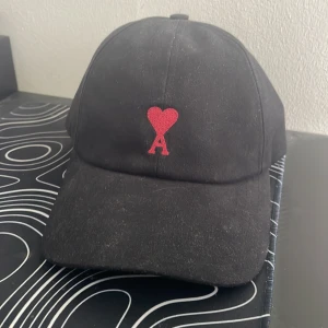 Svart keps med rött hjärta från Ami Paris - Svart keps i bomull med röd broderad logga i form av ett hjärta och bokstaven A framtill. Justerbar rem baktill i metall. Enkel och stilren design som passar till det mesta.