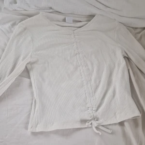 Vit ribbad långärmad topp med knyt - Säljer en vit ribbad långärmad topp med v-ringning och knyt framtill. Toppen har en lätt croppad passform och är perfekt för en stilren vardagslook.