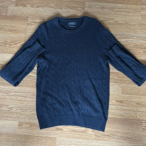 Mörkblå stickad tröja från East West - Mörkblå stickad tröja från East West med klassiskt kabelstickat mönster. Tröjan har rund halsringning, långa ärmar och ribbade muddar vid ärmslut och nederkant. Perfekt för kyligare dagar.