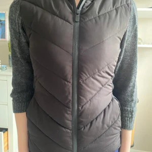 Svart dunväst med luva - Svart dunväst med dragkedja framtill och luva. Västen har ett stilrent, quiltat mönster och är något längre i modellen. Perfekt för lager-på-lager och passar till många olika outfits.