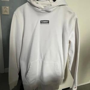 Vit hoodie från Tommy Jeans - Säljer en stilren vit hoodie från Tommy Jeans med logga på bröstet och ärmen. Tröjan har huva med dragsko och en stor magficka. Perfekt för dig som gillar klassisk och enkel streetwear.