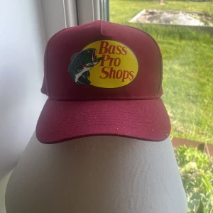 Vinröd keps från Bass Pro Shops - Säljer en vinröd keps med gult och rött Bass Pro Shops-tryck framtill och en fiskgrafik. Kepsen har böjd skärm och mesh-paneler bak för extra ventilation. Perfekt för utomhusaktiviteter och har justerbar passform.