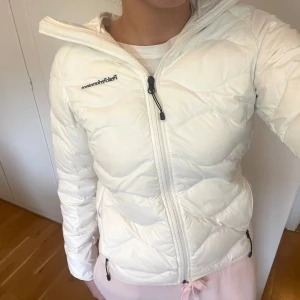 Vit dunjacka med luva från Peak Performance - Säljer en vit dunjacka från Peak Performance. Jackan har en luva, dragkedja framtill och två sidofickor med dragkedja. Lätt och smidig design, perfekt för kyliga dagar! Storlek XS ENDAST använd ett par få gånger, den har en liten liten rosa sminkflävk från en läpppenna förmodligen - vid ena fickan.❣️skicka gärna ett meddelande vid fler frågor. 
