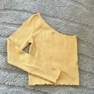 Gul ribbad oneshoulder topp - Trendig gul oneshoulder topp med lång ärm och ribbad struktur. Perfekt för dig som vill sticka ut med en cool och modern stil. Avslutet nertill har en vågig kant för extra detalj.