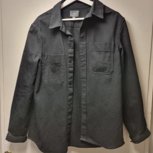 Svart overshirt från H&M - Stilren svart overshirt från H&M med två bröstfickor och knäppning framtill. Skjortan har klassisk krage och långa ärmar, perfekt att bära som lager över en t-shirt eller hoddie.