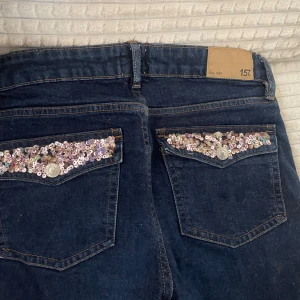 Mörkblå jeans med paljettdetaljer från 157 - Mörkblå jeans från 157 med raka ben och snygga paljettdetaljer i silver och lila på bakfickorna, som jag sytt på själv! Klassisk femficksmodell med dragkedja och knapp. Perfekta för dig som vill ha något extra på dina jeans!Pris kan diskuteras och köparen står för frakten💕