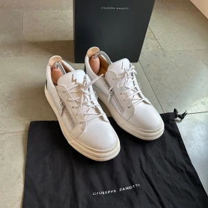 Vita sneakers från Giuseppe Zanotti med svarta detaljer - Säljer ett par vita sneakers från Giuseppe Zanotti med svarta hälpartier och silverfärgade dragkedjor på sidorna. Skorna har rund tå, platt sula och snörning framtill. Ikonisk blixtdetalj på plösen. Perfekta för en stilren look. Skick 9/10 nästa helt oandvända allt OG följer med nypris runt 6000kr. Bara skriva vid funderingar, prisförslag och fler bilder 