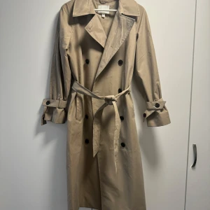 Beige trenchcoat med bälte - Säljer min trenchcoat som köptes nyligen och endast använd ca 3 gånger. Finns dock en liten fläck som inte går bort, annars mycket bra skick!