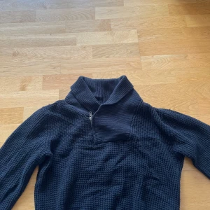 Svart stickad tröja med dragkedja från Bläck - Säljer en svart stickad tröja från Bläck med halv dragkedja och hög krage. Tröjan har ett grovt stickat mönster och ribbade detaljer vid kragen. Perfekt för kyliga dagar.