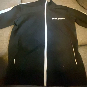 Svart track jacket från Palm Angels - Säljer en svart track jacket från Palm Angels med vit dragkedja och vita ränder längs ärmarna. Jackan har hög krage och logga på bröstet. Perfekt för en sportig och trendig look.