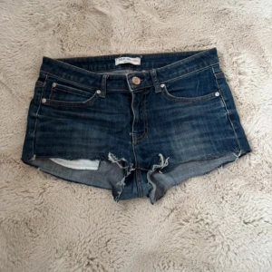 Låg midjade jeansshorts - Snygga mörkblå jeansshorts från Lindex med råa, fransiga kanter och klassisk femficksdesign. Shortsen har låg midja och en avslappnad passform, perfekta för varma sommardagar.