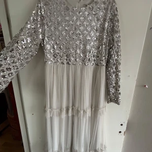 Vit långklänning med paljetter från Reference - Säljer en vit långklänning från Reference med glittriga paljetter på överdelen och långa ärmar. Kjolen är i flera lager av tunn, transparent tyg med volangdetaljer. Perfekt för dig som vill sticka ut med en elegant look.