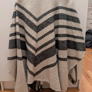 Beige och svart randig halsduk - Stor och mysig halsduk i beige och svart med breda ränder och chevronmönster. Perfekt att svepa runt sig för extra värme. Tjockt och mjukt material som känns skönt mot huden.
